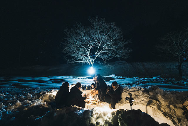 青くライトアップされた夜の雪原の中で料理やお酒を楽しんでいる